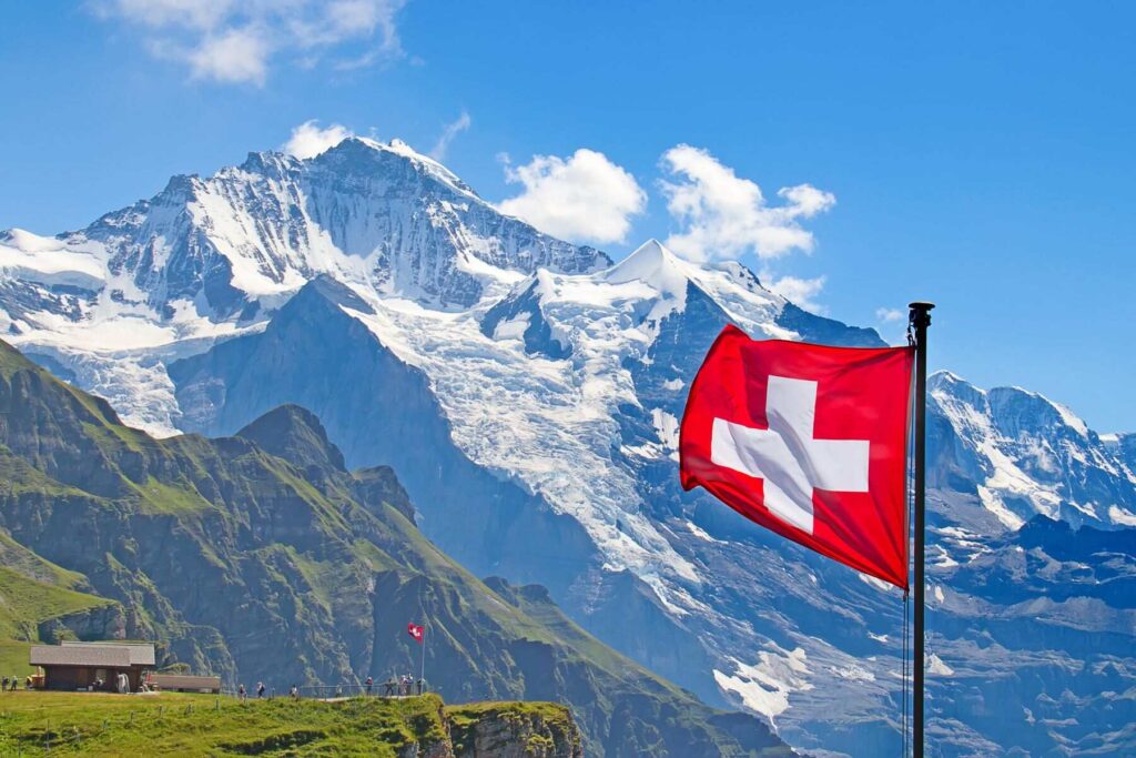 A photo of the Swiss flag, and in the background, an incredible view of the snow-capped Alps and a green hill with tourists standing on it