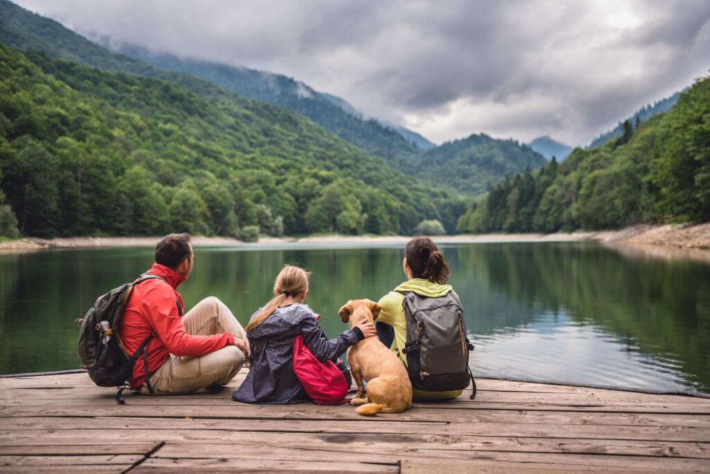 Beautiful photo of a family with a child and a dog sitting on a pier near a lake, with a mountain and dense forest in the background, with backpacks on their backs