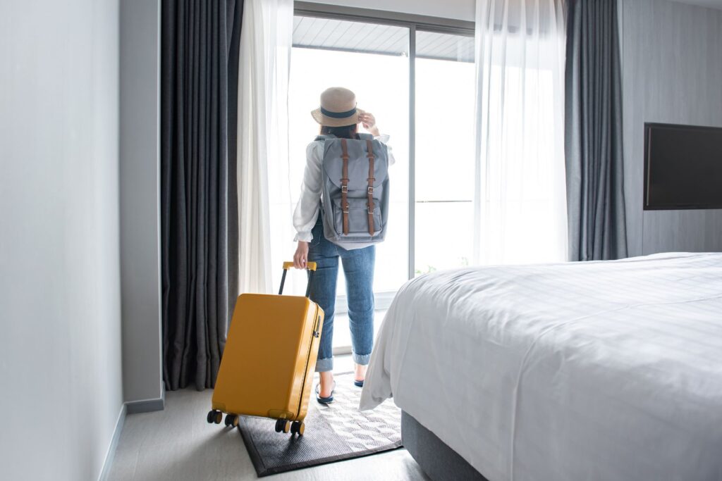 photo of a girl traveling solo in a hotel room standing near the window holding a suitcase with a backpack on her back